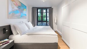 A modern bedroom with a large bed, a bedside table with books, a window with curtains, and a painting on the wall. - 2-room apartment in Charlottenburg-Wilmersdorf (Berlin)