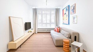 A modern living room with a sofa, a TV, a bookshelf, and a window with curtains. - 2-room apartment in Charlottenburg-Wilmersdorf (Berlin)