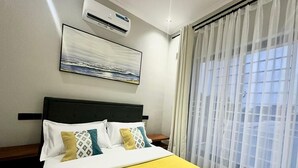 Una habitación de hotel con cama, mesita de noche, aire acondicionado, ventana con cortinas translúcidas y un cuadro en la pared.
