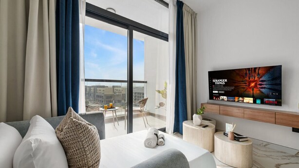 Een moderne hotelkamer met een grote televisie waarop 'Stranger Things' wordt weergegeven, een balkon met uitzicht op de stad, en een keurig opgemaakte zithoek.