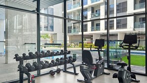 Een sportschool met trainingsapparatuur, grote ramen en uitzicht op een gebouw en groen.