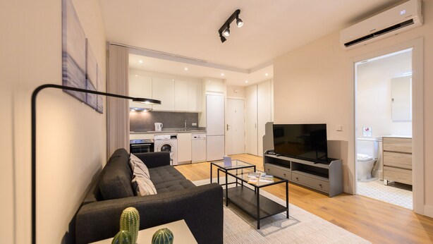 A modern living room with a sofa, coffee table, and TV. There is a kitchen in the background with cabinets and appliances. - Bright one-bedroom flat with excellent amenities (Madrid)