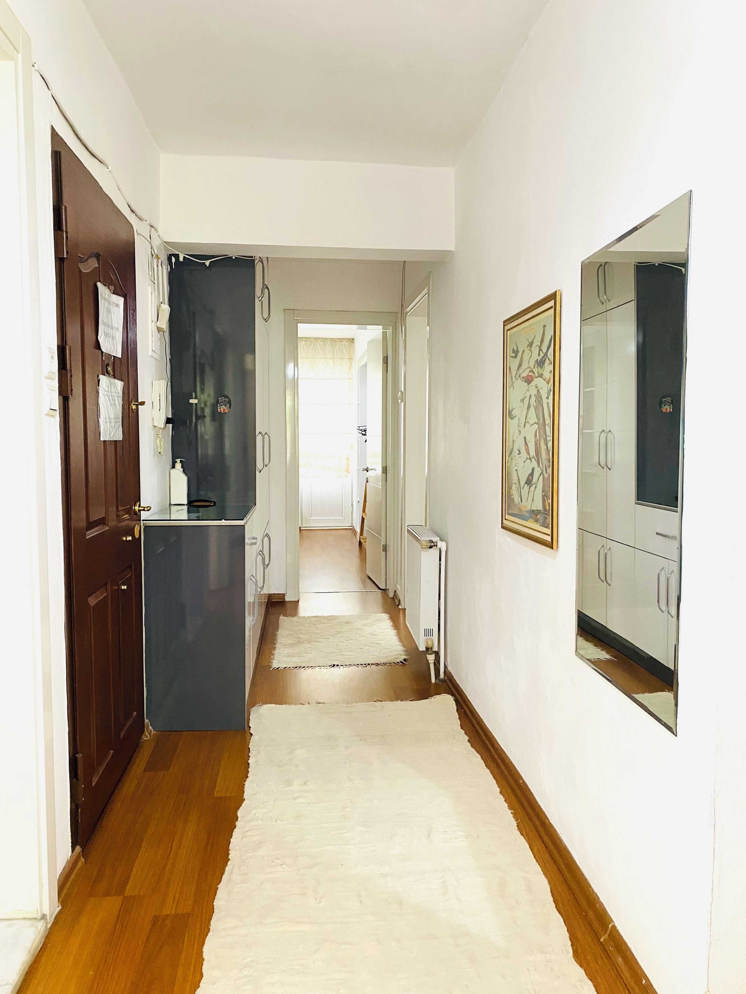 A narrow hallway with wooden flooring, a white rug, and a kitchen area with a refrigerator and cabinets.