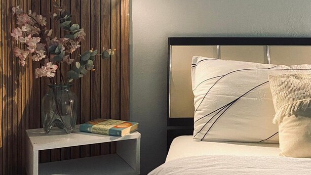 A bedroom with a wooden panel wall, a bedside table with a vase of flowers, a bed with white bedding, and a framed picture on the wall. - Aydinoglu Suites (Denizli)
