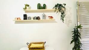 A room with a bed, a wall-mounted shelf with decorative items, a hanging plant, and a window with blinds. - Aydinoglu Suites (Denizli)