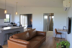 Een moderne woonkamer met een bruine leren bank, een eethoek met een tafel en stoelen, een keuken met een bar en een groot raam dat zorgt voor veel daglicht.