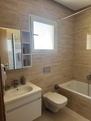 Ein modernes Badezimmer mit einer Badewanne, Toilette und Waschbecken. Über dem Waschbecken befindet sich ein Fenster und eine Handtuchstange mit Handtüchern.
