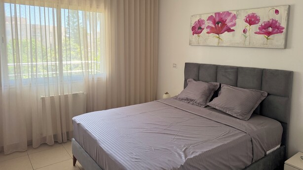A bedroom with a large bed, a window with sheer curtains, and a floral painting on the wall.