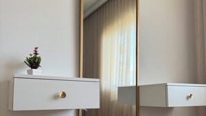 A modern bathroom with a large mirror, two white cabinets with gold handles, a small plant on the left cabinet, and a round stool with a gold base.