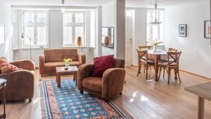 Un vaste espace de vie comprenant un canapé, des fauteuils, une table basse et une table à manger avec des chaises. De grandes fenêtres laissent entrer la lumière naturelle, un tapis à motifs recouvre le sol et un luminaire est fixé au plafond.