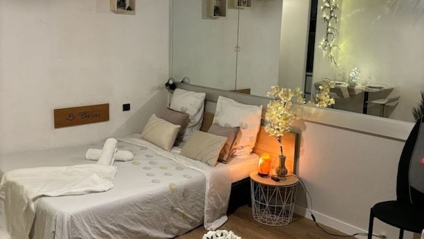 Un dormitorio con una cama, una mesita de noche con lámpara, un espejo y una pequeña estantería decorada.