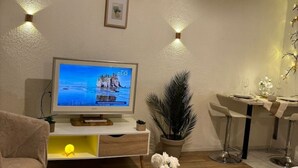 Una sala de estar con un televisor que muestra una imagen panorámica, una zona de comedor con una mesa dispuesta para dos personas y una planta en maceta.