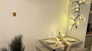 Un comedor con una mesa dispuesta para dos personas, dos sillas, una pequeña planta en maceta y una lámpara fijada a la pared.