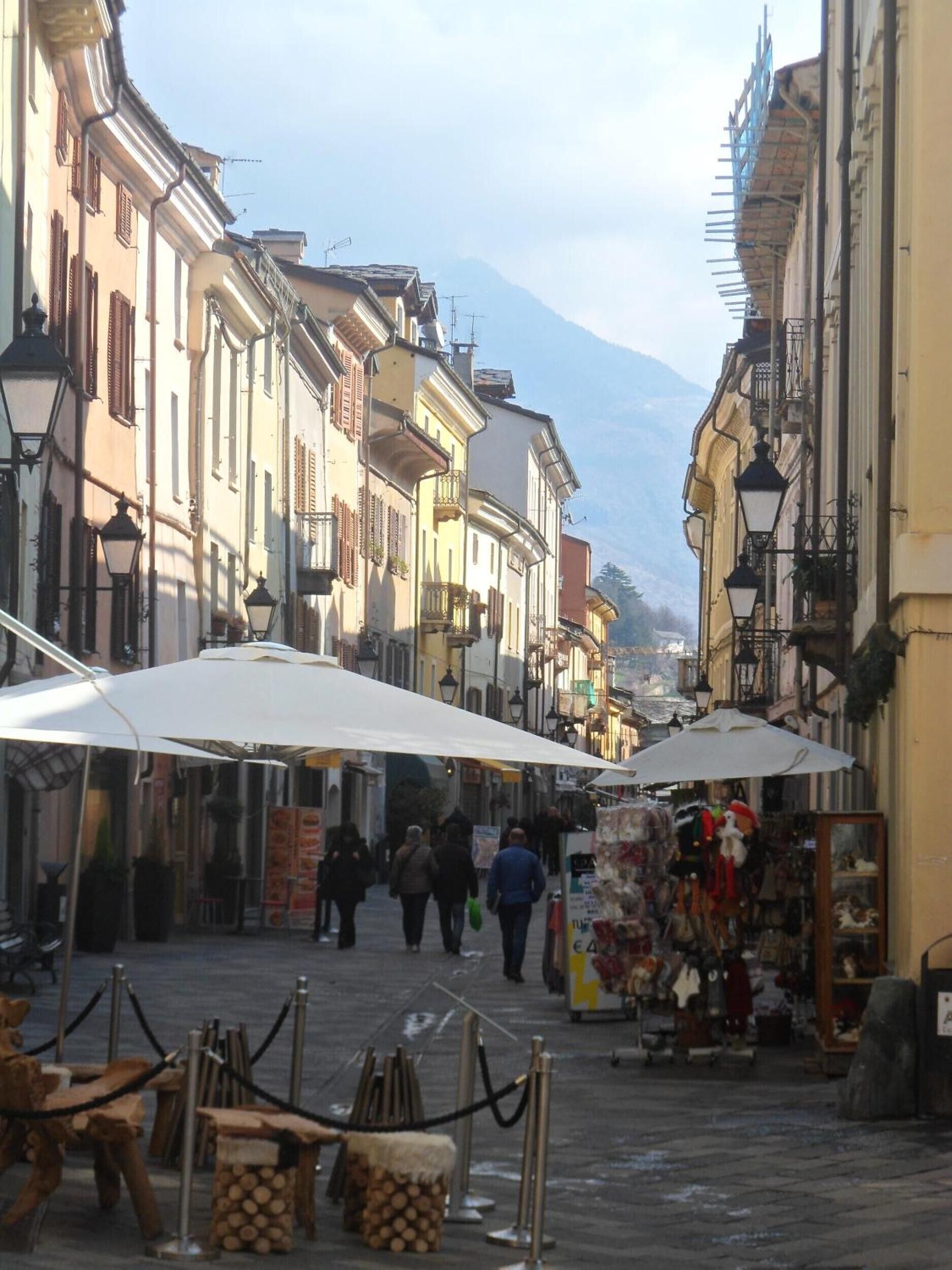 A narrow street with buildings on either side, outdoor seating, and a mountain in the background.