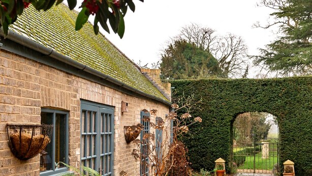 A brick building with a tiled pathway leading to a gate, surrounded by potted plants and a hedge. - The Potting Shed at Island Hall (Godmanchester)