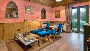 A cozy room with wooden walls, a large window, and a view of the garden. - The Potting Shed at Island Hall (Godmanchester)