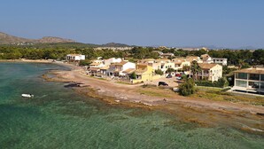 A coastal town with buildings along the shore, clear water, and a hilly background. - Alcudia 1 | Seafront Barcares Villa | Frontline sea views and spacious 6-bed. (Pollensa)
