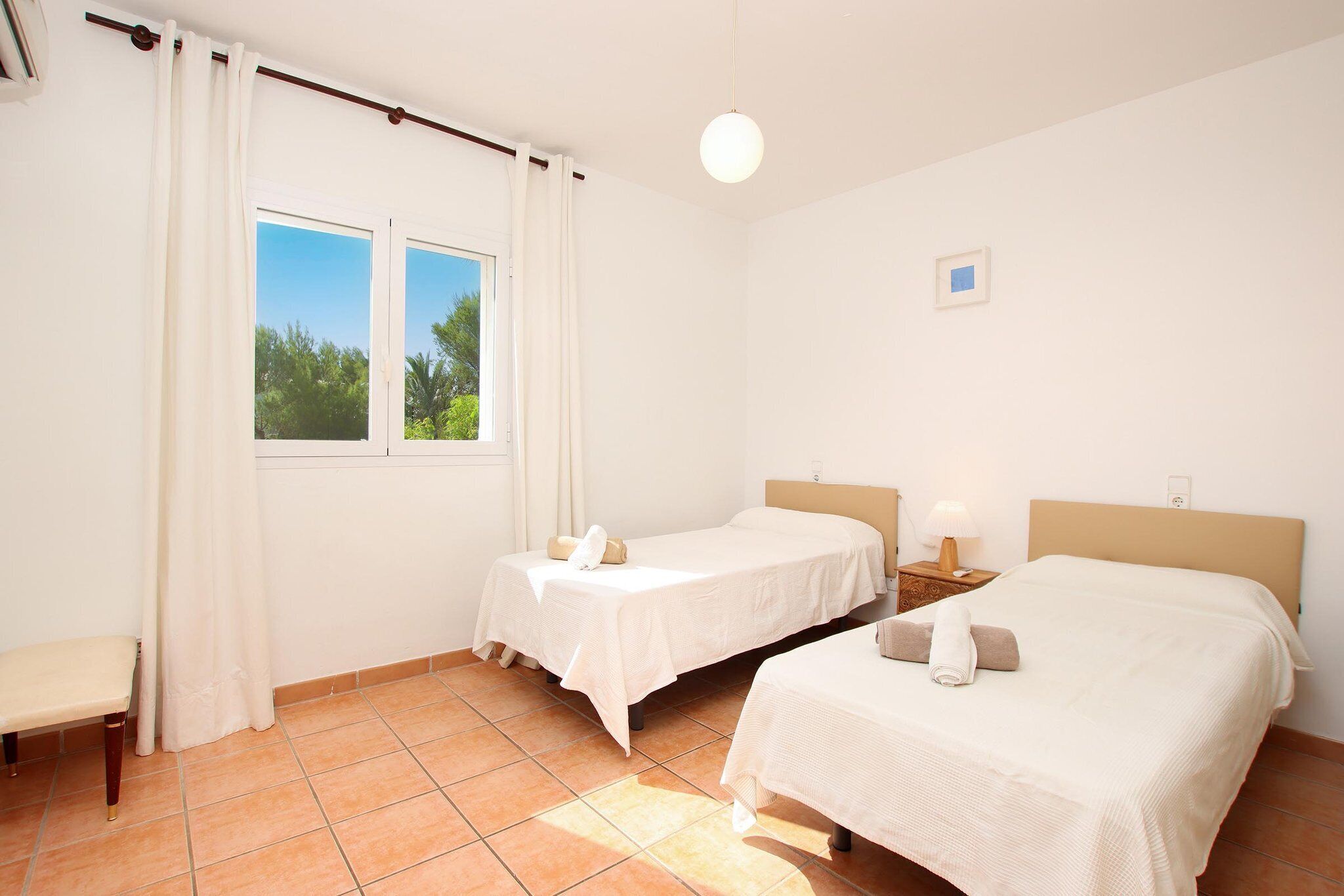 Une chambre d’hôtel avec deux lits simples, un banc, une petite table et une fenêtre avec des rideaux.