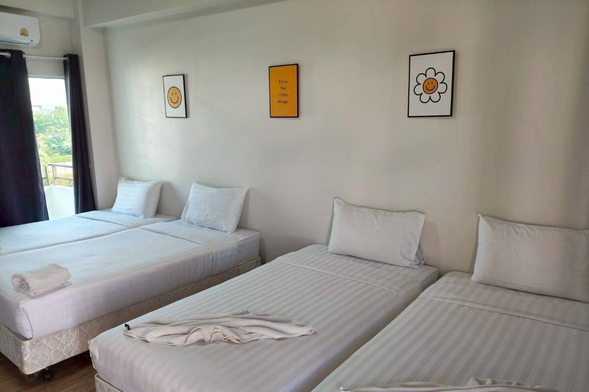 Quarto de hotel com duas camas individuais, cada uma com uma almofada e uma toalha dobrada. Há duas imagens emolduradas na parede, uma com uma carinha sorridente e outra com uma flor.