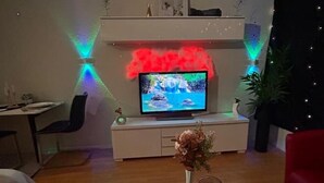 Ett modernt vardagsrum med en platt-TV placerad på en vit TV-bänk, en matplats med ett bord och stolar samt en dekorativ vas med torkade blommor på ett sidobord.