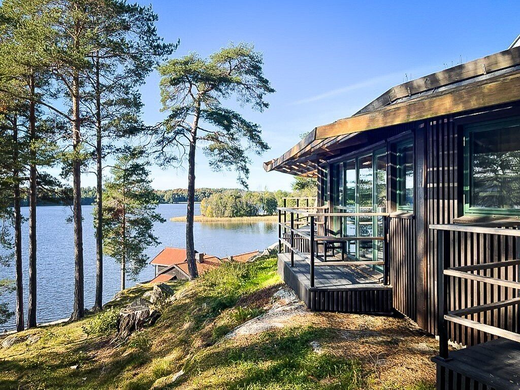 En stuga vid sjön med en träveranda och stora fönster med utsikt över vattnet.