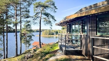 Järven rannalla sijaitseva mökki, jossa on puinen terassi ja suuret ikkunat, joista avautuu näkymä vedelle.