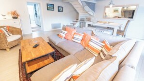 A modern living room with a sofa, coffee table, and wicker chair. A staircase leads to a dining area with a table and chairs. - Charming farmhouse in the town center and within walking distance of the beaches (Larmor-Plage)