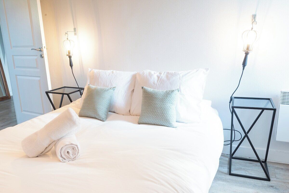 Una cama bien tendida con sábanas blancas y dos almohadas, una puerta blanca y dos mesitas de noche negras con lámparas fijadas a la pared.