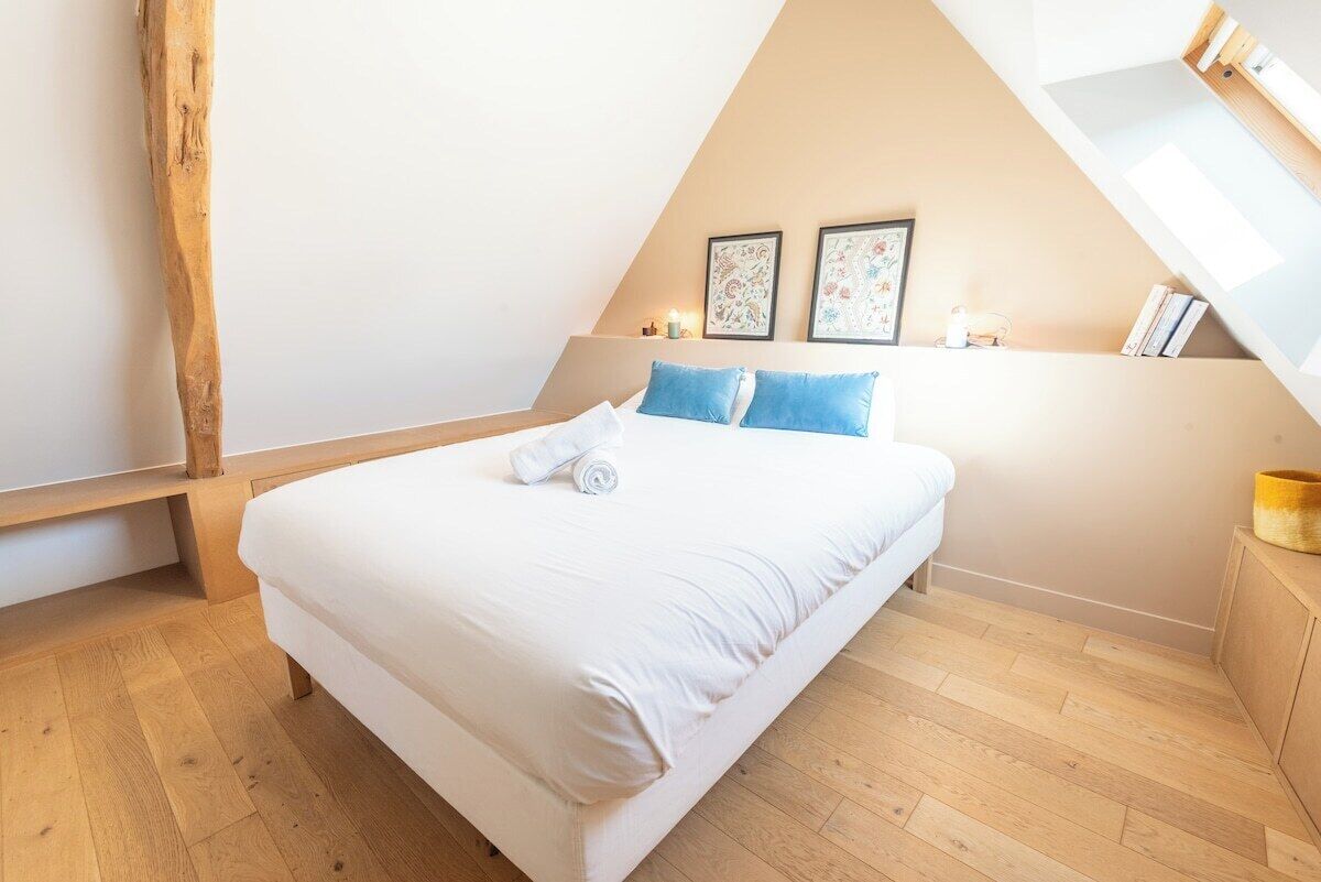 A bedroom with a wooden floor, a bed with white bedding and blue pillows, a bench, and two framed pictures on the wall.