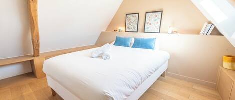 A bedroom with a wooden floor, a bed with white bedding and blue pillows, a bench, and two framed pictures on the wall.