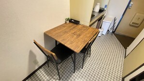 Ein kleiner Essbereich mit einem Holztisch und zwei Stühlen. Auf dem Tisch steht ein kleiner Topfpflanze, daneben befindet sich ein Mülleimer. Der Boden ist mit einem gemusterten Fliesenbelag versehen.