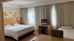 A hotel room with a large bed, a television on a wooden stand, a desk with a chair, and a window with curtains. - Guarulhos Flats Services (Guarulhos)