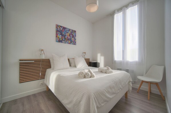 Een moderne slaapkamer met een groot bed, een nachtkastje, een stoel, een kleurrijk abstract schilderij en een raam met lichte gordijnen.