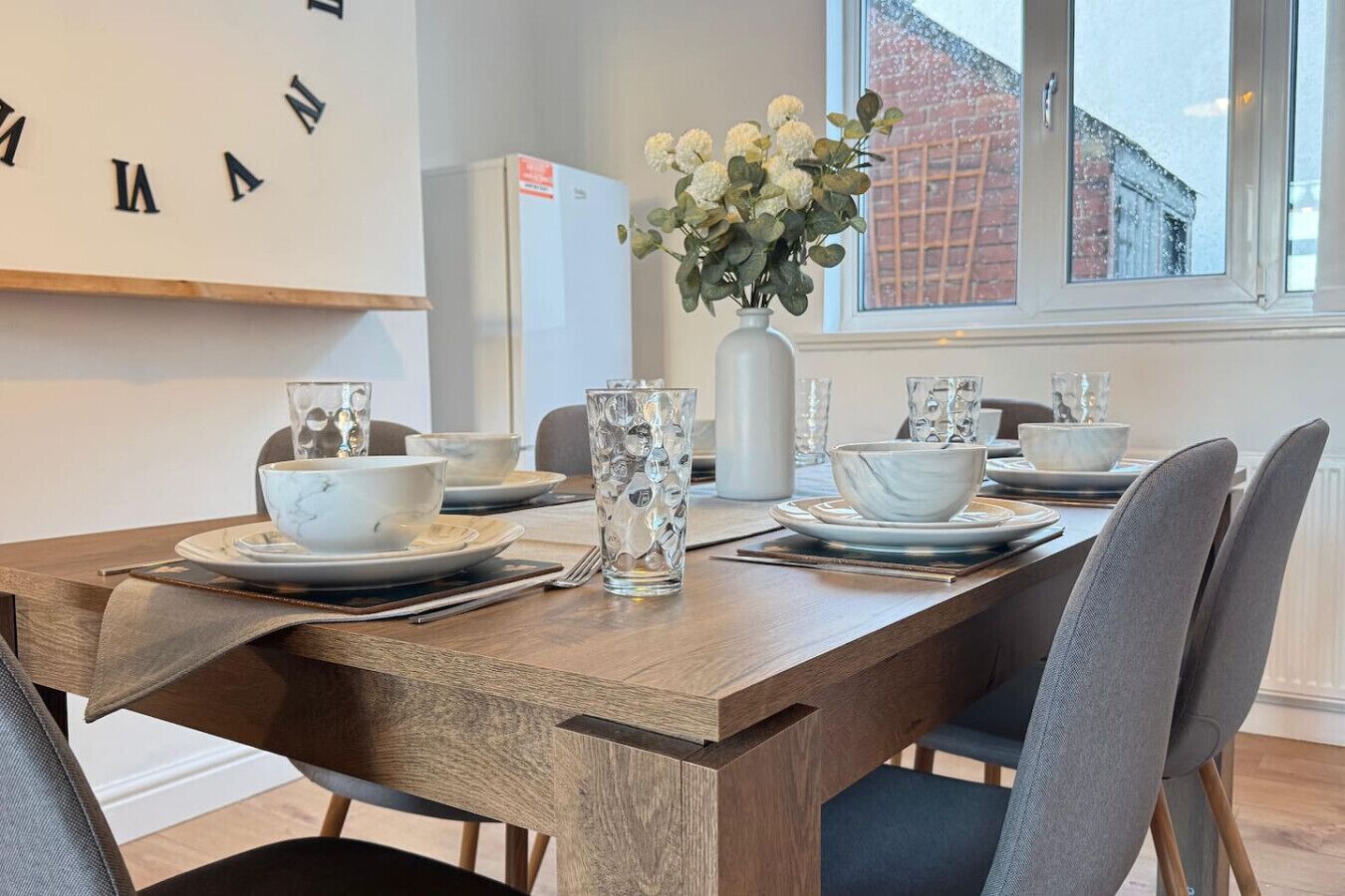 A dining area with a wooden table set for four, including bowls, glasses, and a vase with flowers.