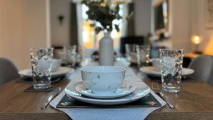 A dining table set with plates, bowls, glasses, and cutlery, centered in a room with a vase of flowers in the background.