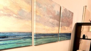 Een beige bank met kussens, een houten bijzettafeltje en drie ingelijste schilderijen van een zeegezicht aan de muur.