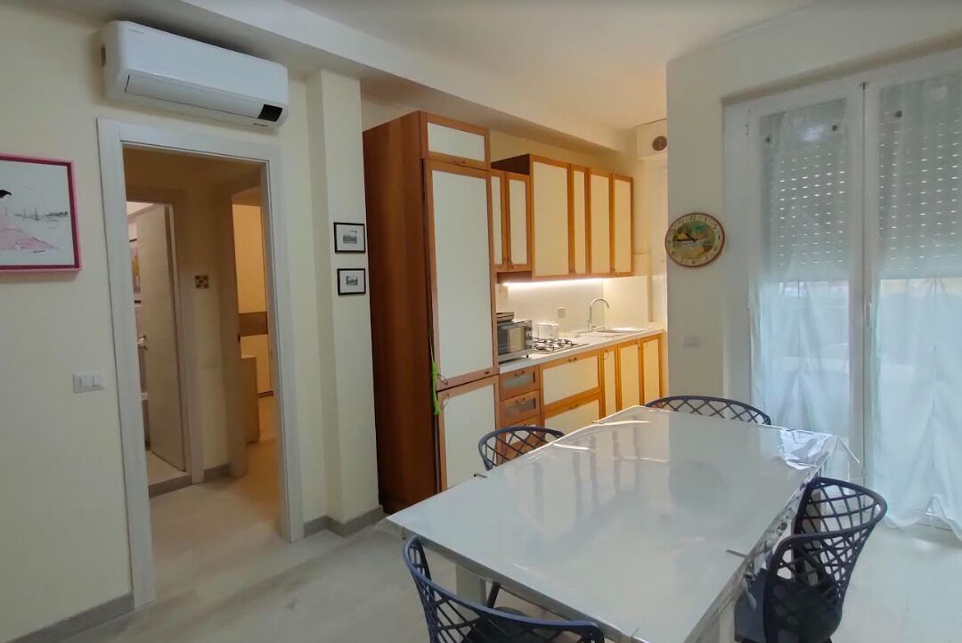 A modern kitchen with wooden cabinets, a dining table with chairs, and a view of the outside through a window with curtains.