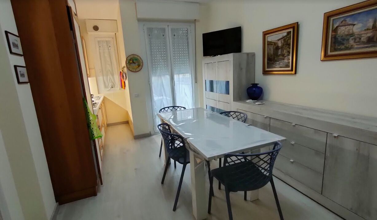 A dining area with a white table and chairs, a TV mounted on the wall, and a cabinet with a vase.