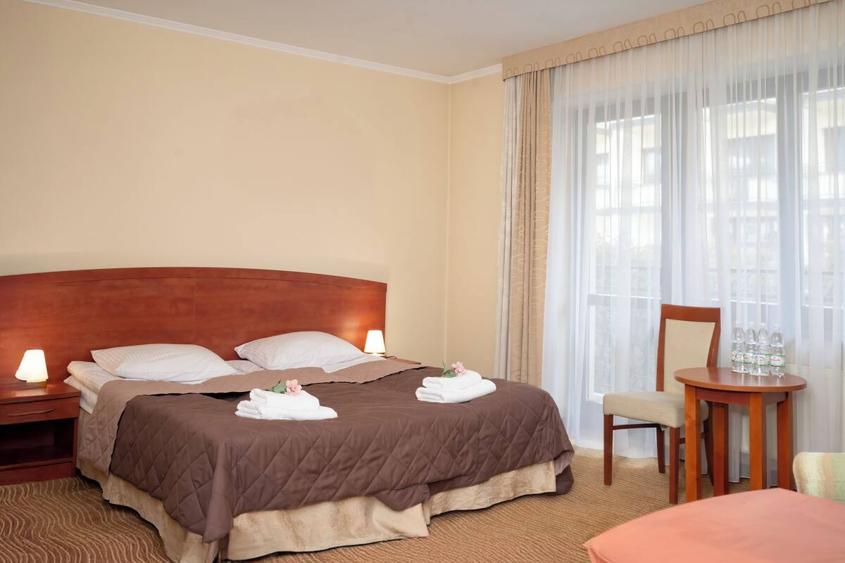 Camera d'albergo con un letto grande, una testiera in legno, un comodino con una lampada, una sedia e un tavolino con bottiglie.