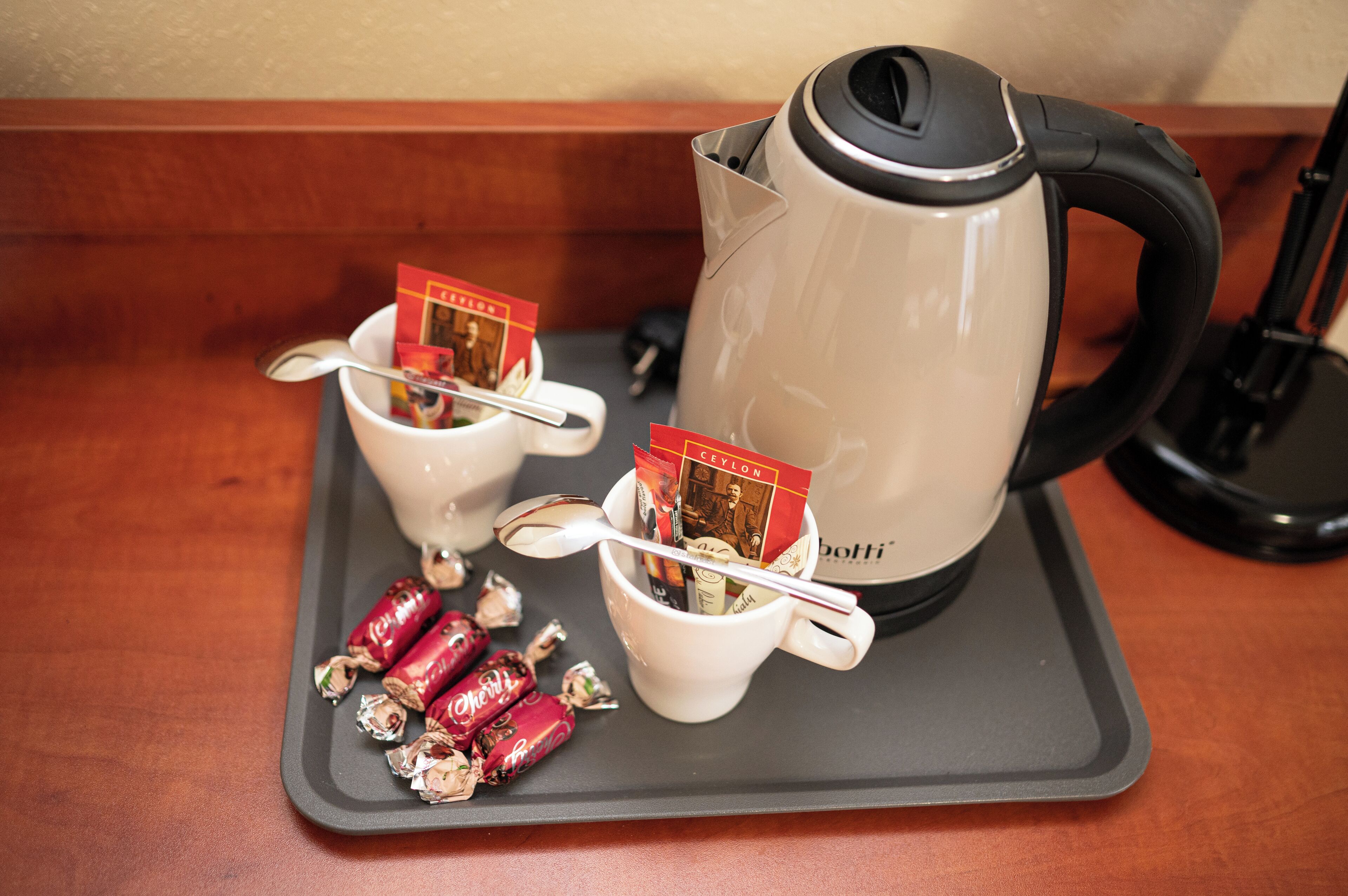 A tray with two white mugs, each containing a spoon and a card, and a box of chocolates. A beige electric kettle is also visible.