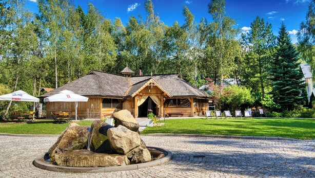 A wooden lodge with a stone fountain, surrounded by trees and a well-maintained lawn.