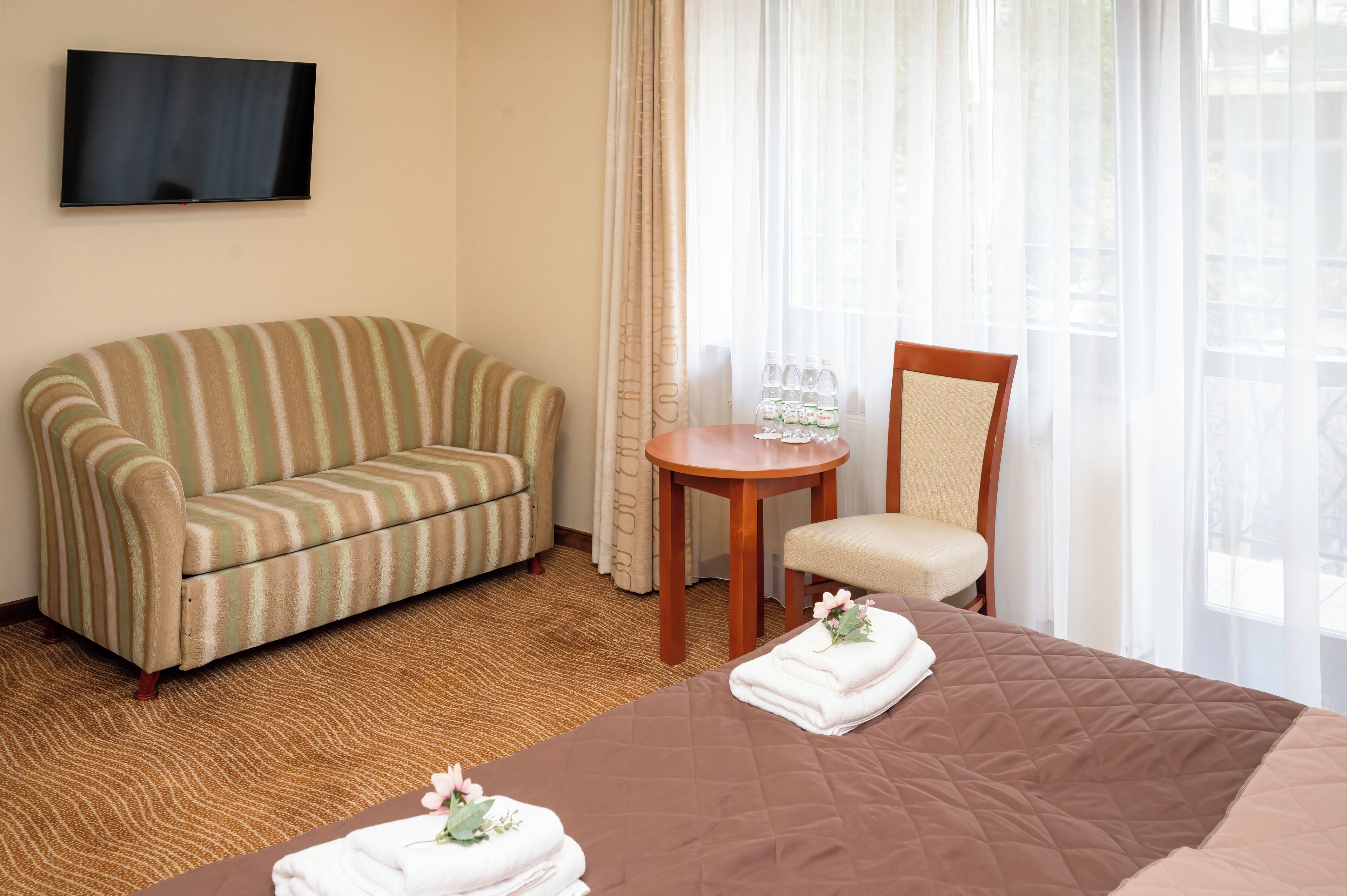 A hotel room with a bed, a striped sofa, a wooden chair, a small round table with water bottles, and a TV mounted on the wall.