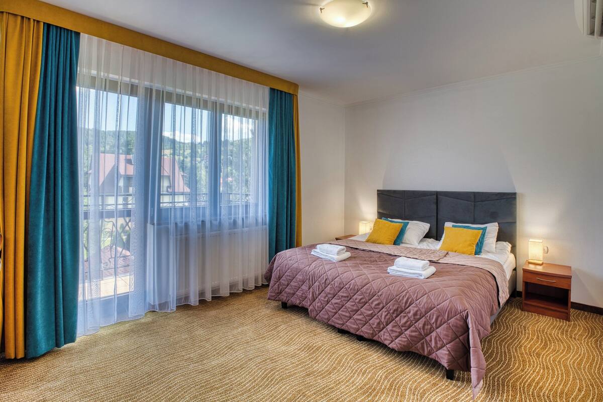 Una camera d'hotel con un letto grande, un comodino e vista all'esterno attraverso tende leggere.