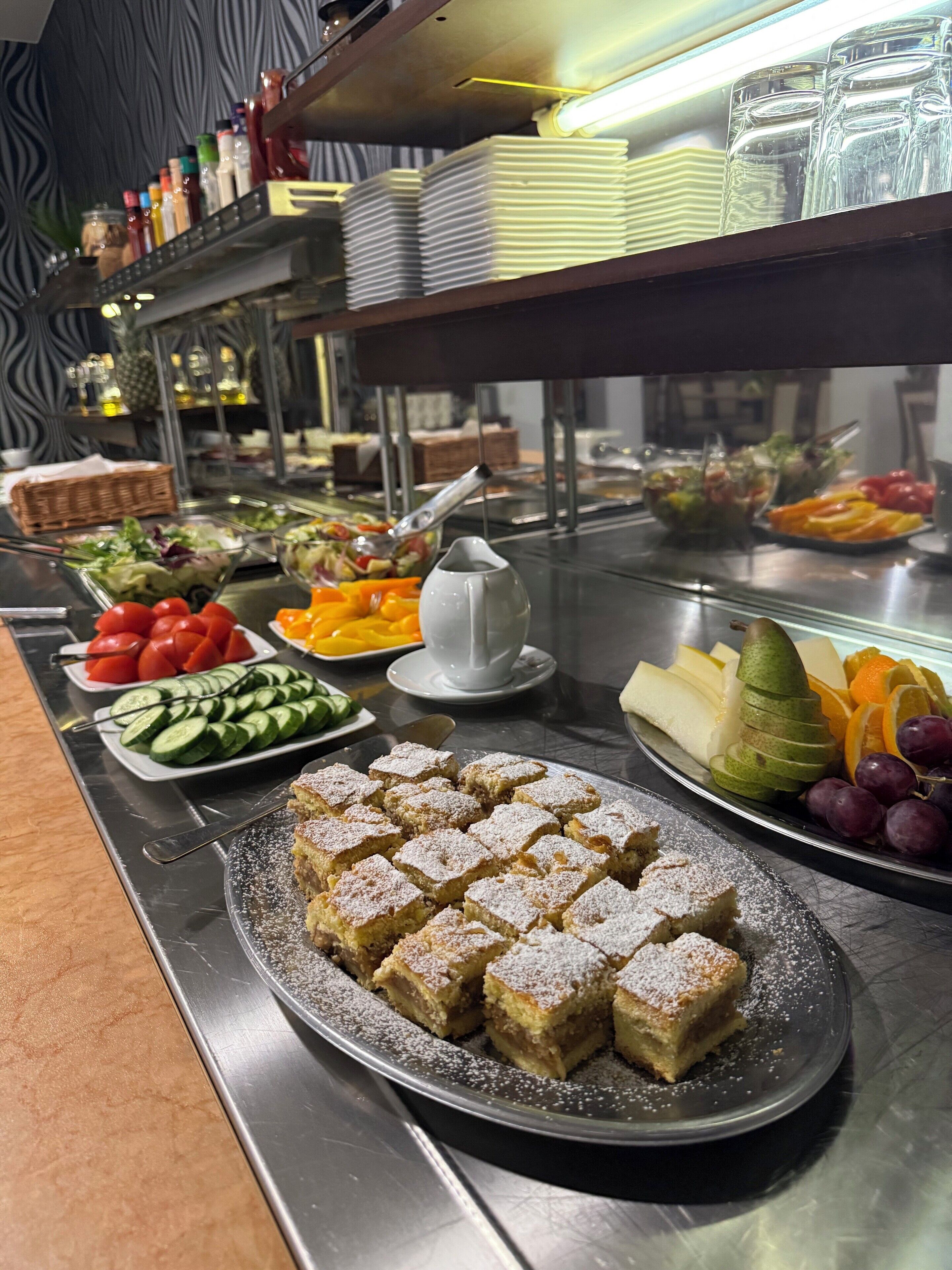 A buffet with various food items including sliced cucumbers, cherry tomatoes, a plate of fruit, and a plate of powdered dessert.