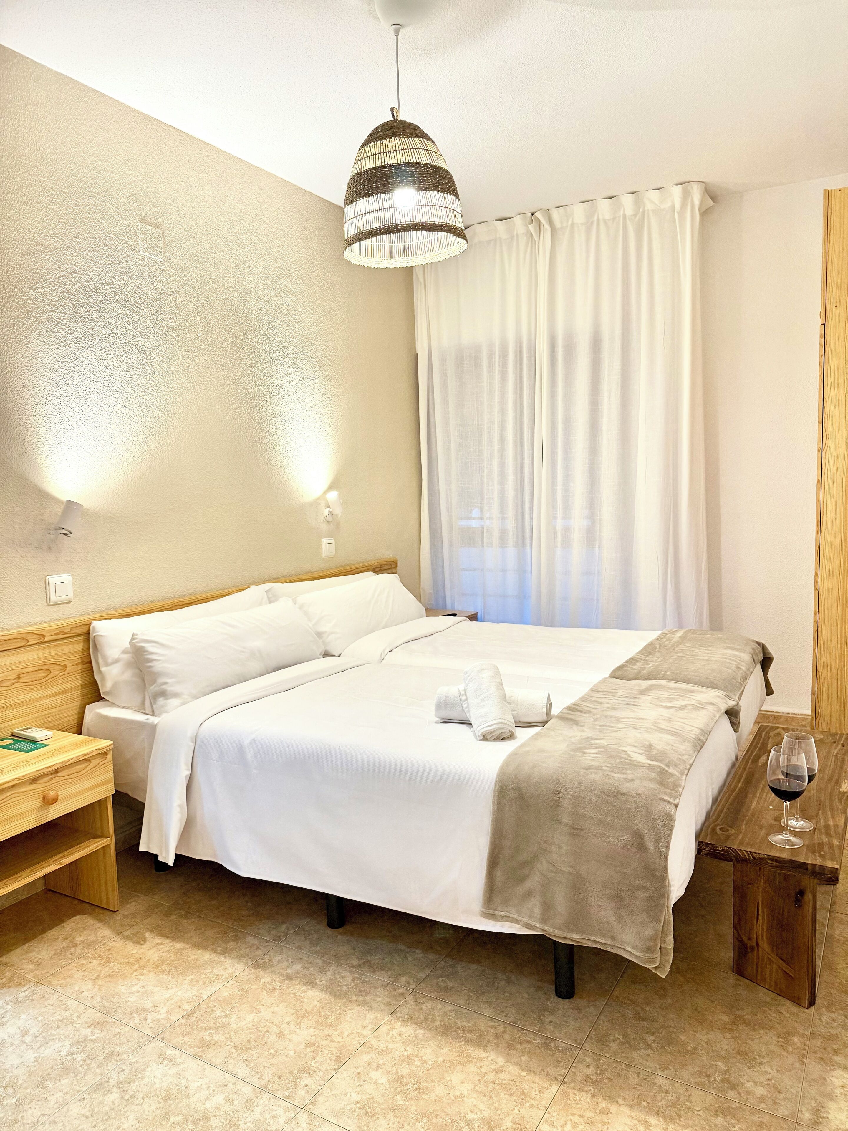 Ein Hotelzimmer mit einem großen Bett, einem Nachttisch mit einem Weinglas, einer Lampe und einem Fenster mit Vorhängen.