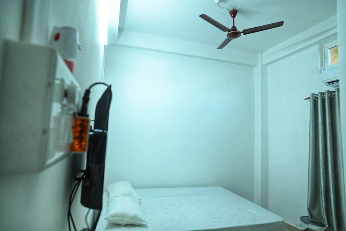 Egy kis, tiszta szoba mennyezeti ventilátorral, ággyal és radiátorral.