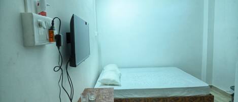 A small, simple room with a bed, a TV mounted on the wall, a small table with a remote and glasses, and a wall socket.