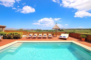 A swimming pool with lounge chairs and a thatched umbrella, surrounded by wooden decking and a view of distant mountains. - Finca Casou | Pollensa | Spacious countryside retreat with pool for. (Pollensa)