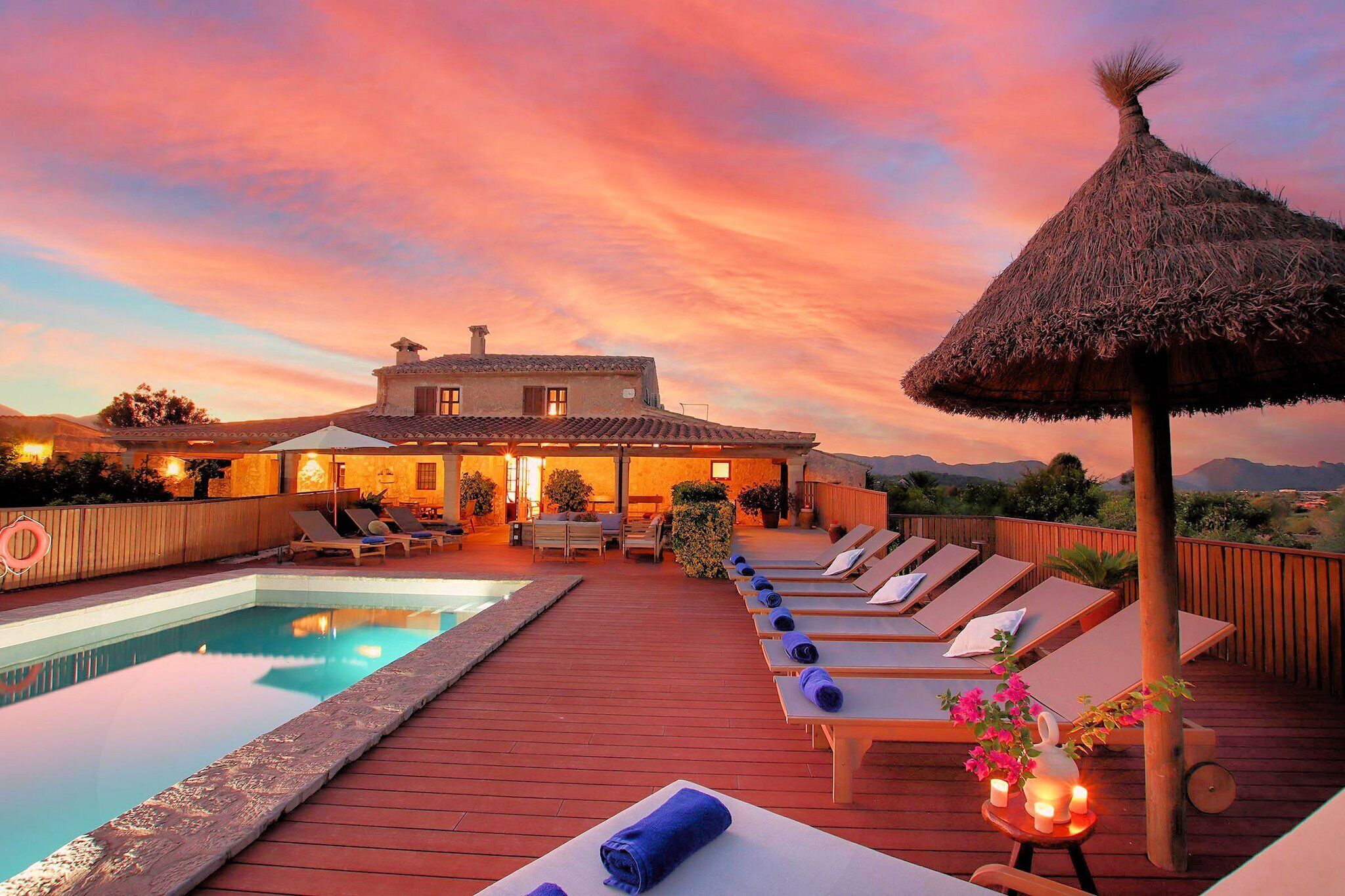 Zona junto a la piscina con sillones de descanso y una sombrilla de paja, una terraza de madera y una casa al fondo.