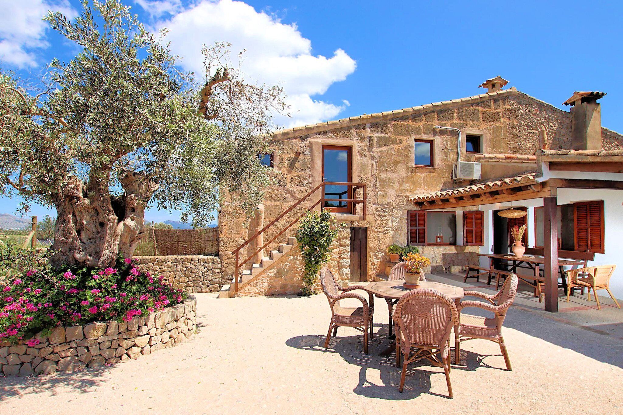 A rustic stone building with a wooden staircase, outdoor seating area, and a large tree with pink flowers.
